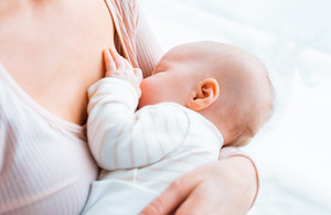 Eine junge Mutter sitzt am Fenster, hält ihr Baby im Arm und stillt