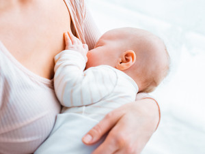 Eine junge Mutter sitzt am Fenster, hält ihr Baby im Arm und stillt