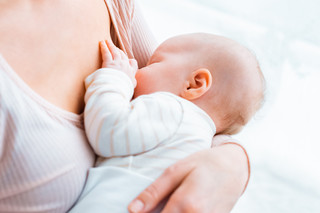 Eine junge Mutter sitzt am Fenster, hält ihr Baby im Arm und stillt