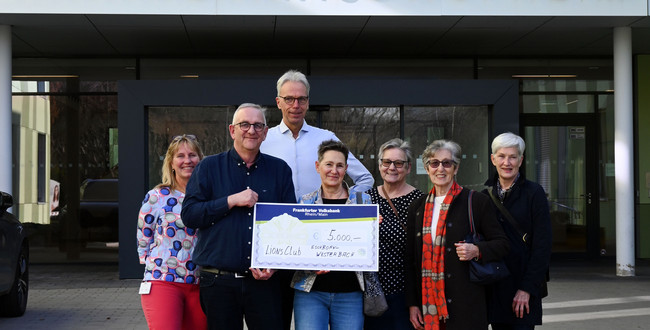 MAn sieht eine Gruppe von Menschen vor dem Krankenhauseingang stehen. Zwei davon halten einen übergroßen symbolischen Scheck in den Händen. Darauf die Spendensumme