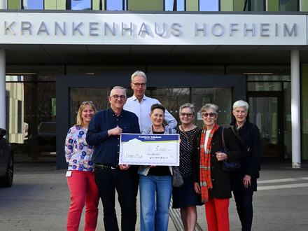 MAn sieht eine Gruppe von Menschen vor dem Krankenhauseingang stehen. Zwei davon halten einen übergroßen symbolischen Scheck in den Händen. Darauf die Spendensumme