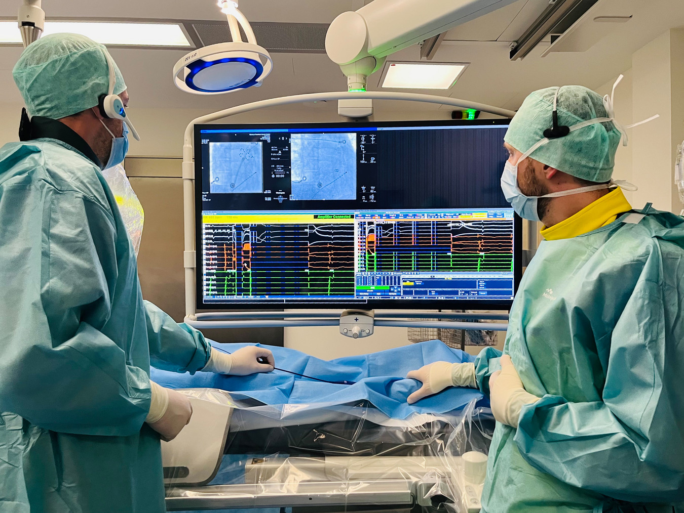 Man sieht zwei Ärzte in blauer OP-Kleidung. Beide schauen auf einen großen Monitor, auf dem das CT-Bild eines Katheters in Gefäßen zu sehen ist, ebenso wie die Darstellung von Messwerten. Sie behandeln einen Patienten. 