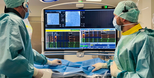 Man sieht zwei Ärzte in blauer OP-Kleidung. Beide schauen auf einen großen Monitor, auf dem das CT-Bild eines Katheters in Gefäßen zu sehen ist, ebenso wie die Darstellung von Messwerten. Sie behandeln einen Patienten. 
