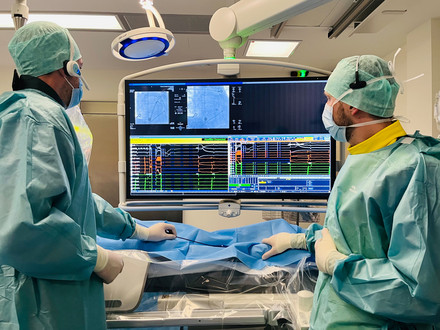 Man sieht zwei Ärzte in blauer OP-Kleidung. Beide schauen auf einen großen Monitor, auf dem das CT-Bild eines Katheters in Gefäßen zu sehen ist, ebenso wie die Darstellung von Messwerten. Sie behandeln einen Patienten. 