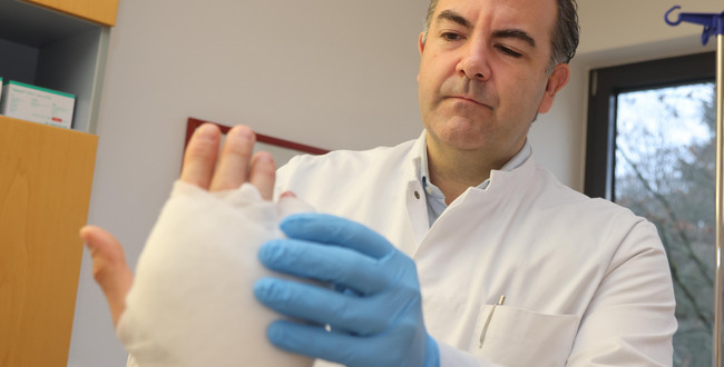 Man sieht den Arzt Dr. Spindler in einem weißen Kittel, wie er eine verbundene Hand untersucht