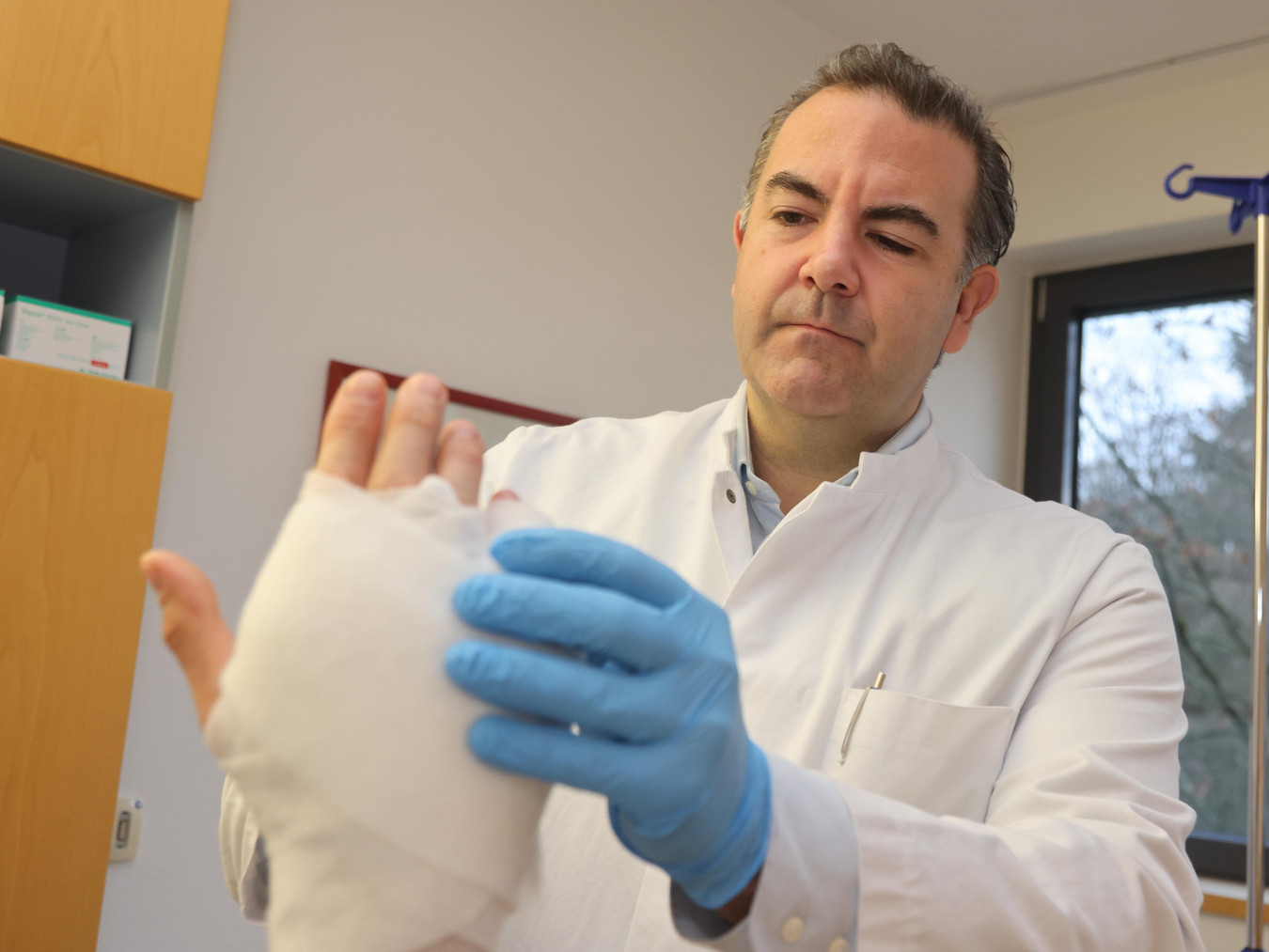 Man sieht den Arzt Dr. Spindler in einem weißen Kittel, wie er eine verbundene Hand untersucht